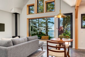 Uma sala de estar moderna com um sofá cinzento, uma mesa de jantar de madeira com cadeiras e amplas janelas com vista para árvores e para o oceano.