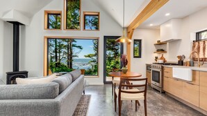 A modern living space with a large window offering a view of the ocean, a cozy seating area, a dining table, and a kitchen with wooden cabinets. - Fogged Inn (Ucluelet)