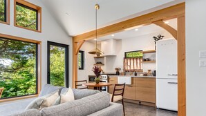 A modern kitchen with wooden beams, a dining area with a gray sofa, and large windows offering a view of greenery. - Fogged Inn (Ucluelet)