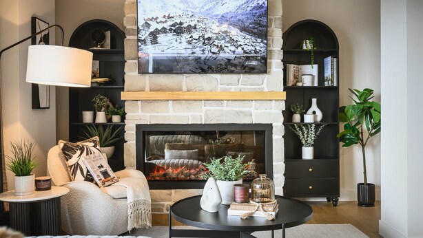 A cozy living room with a stone fireplace, a comfortable armchair, a round coffee table, and a large TV mounted above the fireplace. - Hôtel à la maison - Luxe Alpine MSA (Beaupré)