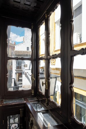 A view through a wooden-framed window with a balcony, showcasing a building with a tiled roof and a sign that reads "ALFONSIN." - CSE Puerta de Armas Apartments (Seville)