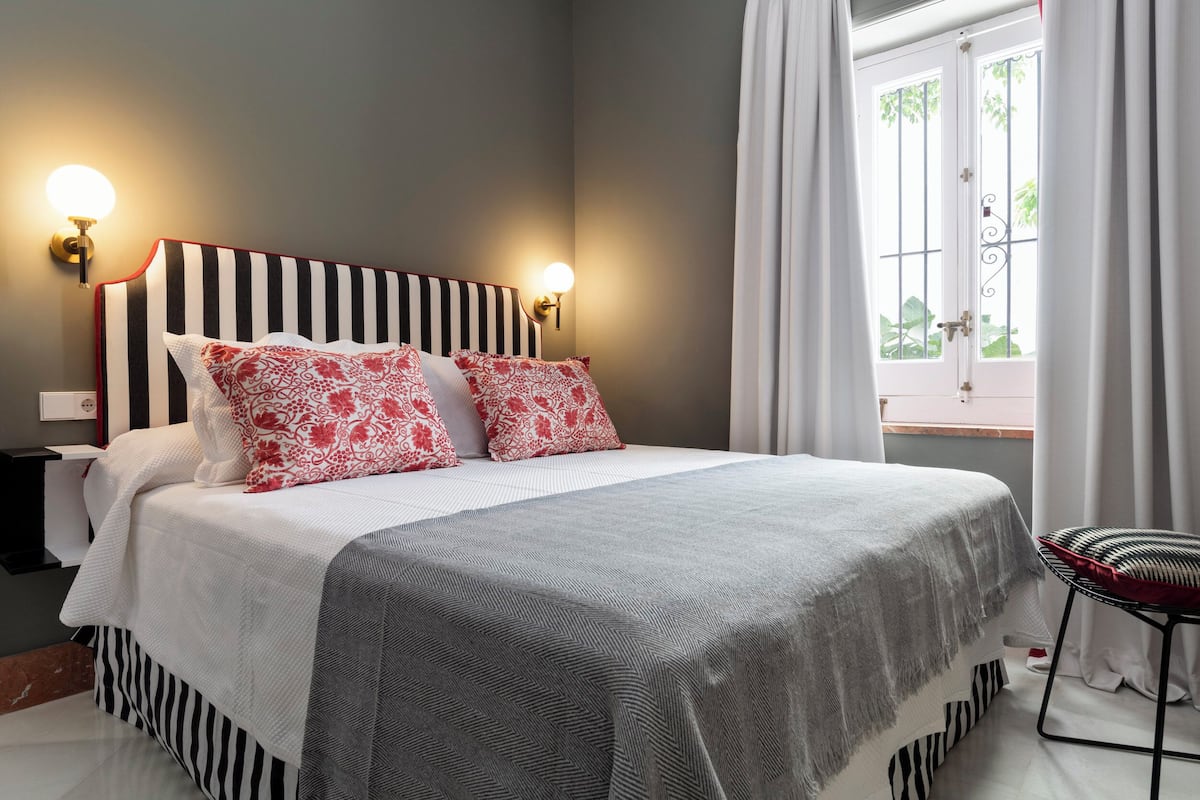 A neatly made bed with striped bedding and decorative pillows, a bedside table with a lamp, and a window with curtains.