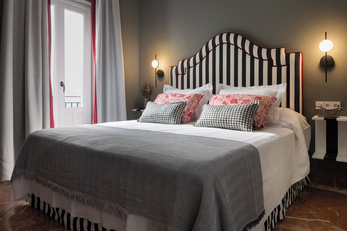 A neatly made bed with striped bedding, a headboard with red trim, and two wall-mounted lights.
