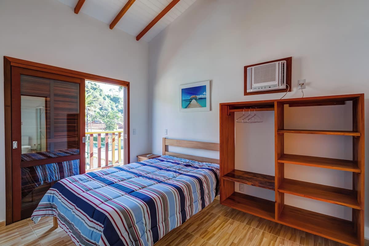 A bedroom with a bed, a wooden wardrobe, a picture on the wall, and a window with a view of greenery.
