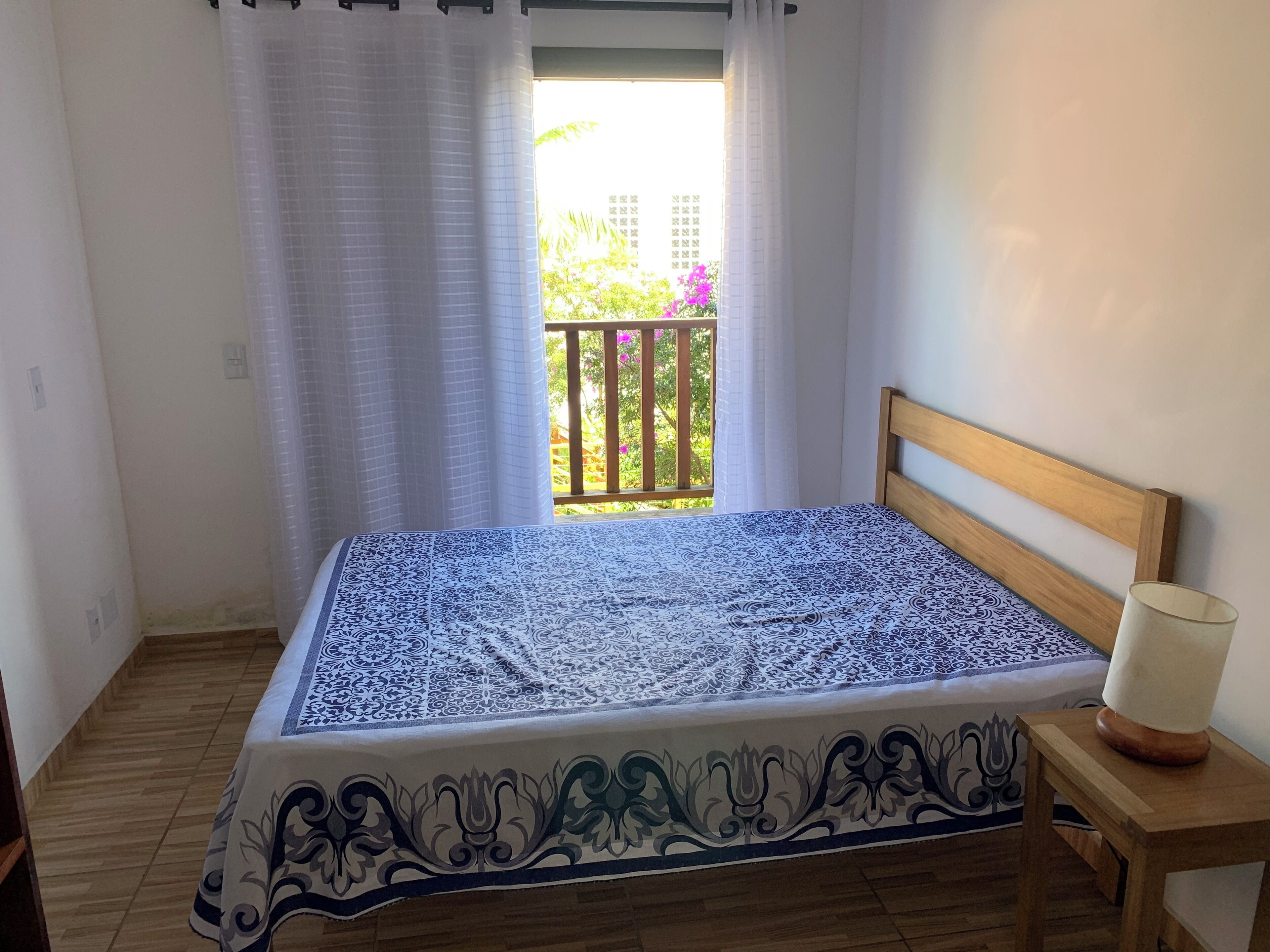 A single bed with a patterned bedspread, a wooden headboard, a bedside table with a lamp, and a window with a view of greenery and a balcony.