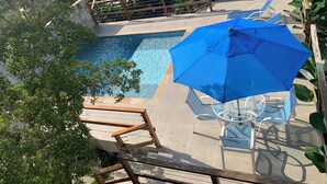 Una piscina con sombrilla azul y sillas de vidrio, rodeada de plantas y una escalera de madera.