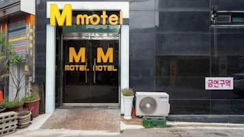Entrada de un motel con un cartel grande con la letra 'M', una placa con el nombre y una unidad de aire acondicionado pequeña.