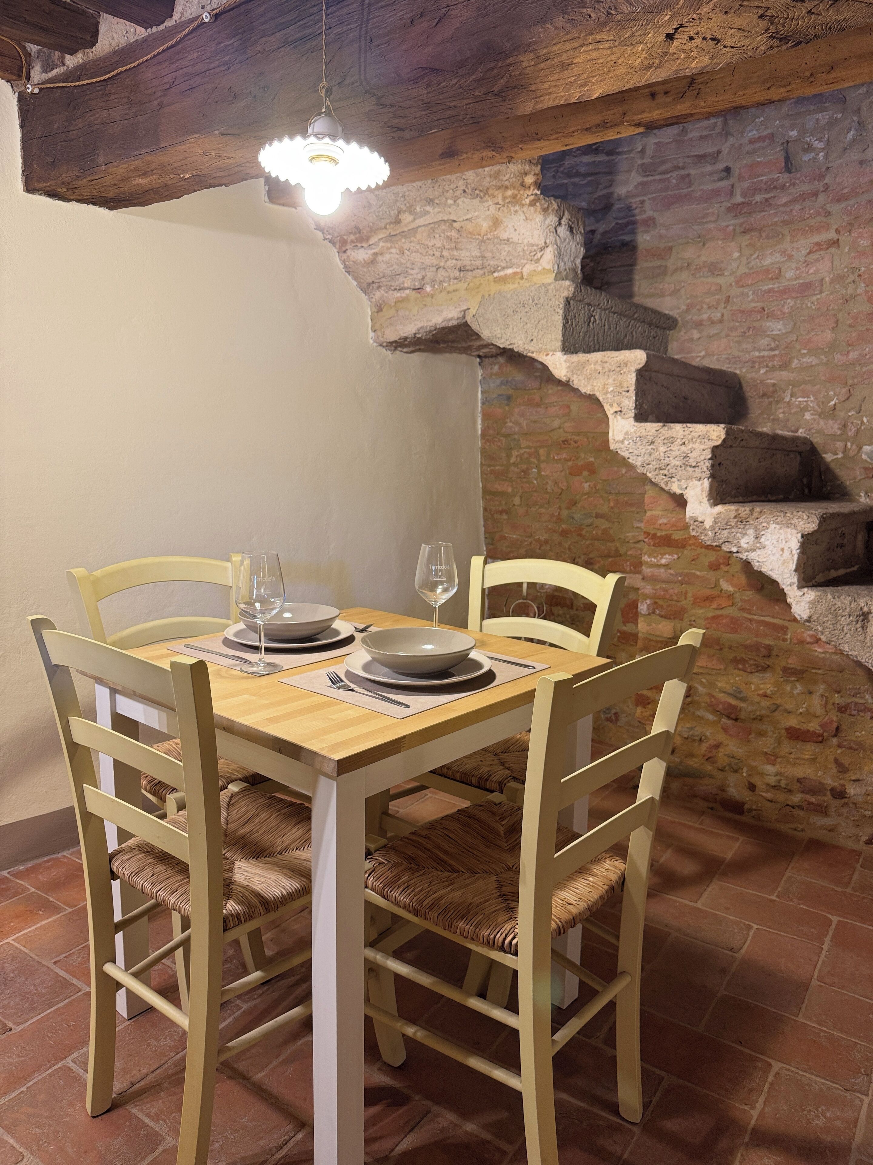 A dining area with a wooden table and chairs, a hanging light fixture, and a brick wall with a stone staircase.