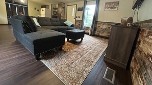 A living room with a sectional sofa, ottoman, patterned rug, ceiling fan, and a TV on a wooden stand.