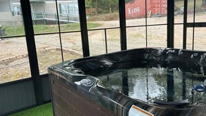 A hot tub with a warning sign, surrounded by a windowed enclosure with a view of a gravel area and a red shipping container.