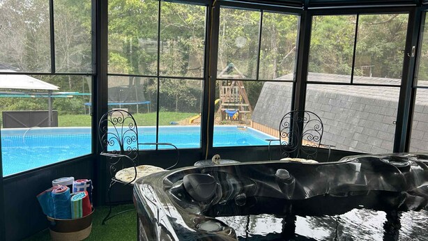 A hot tub in a covered outdoor area with a view of a swimming pool and a wooden play structure in the background.