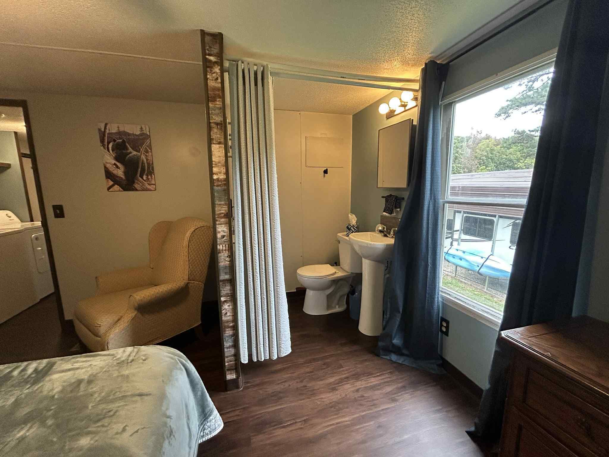 A compact hotel room with a bed, a chair, a toilet, a sink, and a window with a view of a train.