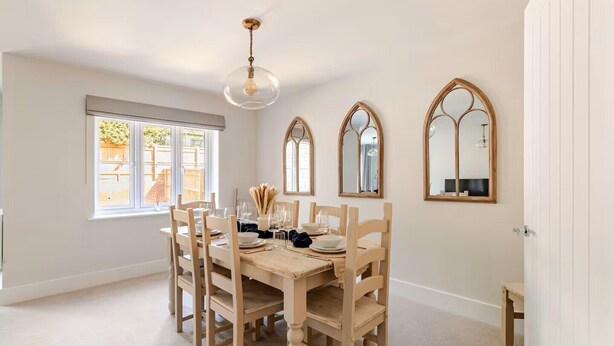 A dining room with a wooden table set for six, three chairs on each side, a hanging light fixture, and three arched mirrors on the wall.