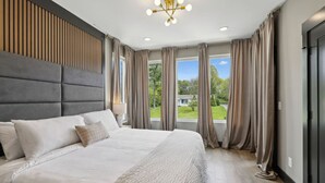 Un dormitorio moderno con una cama grande, un panel de pared de madera, una araña y vistas a un paisaje verde a través de ventanas amplias.