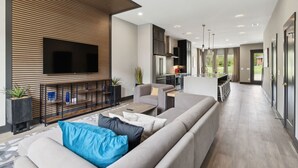 Een moderne woonkamer met een grote flatscreen televisie, een bank met kussens, een salontafel en een keukenhoek met kasten en een koelkast.