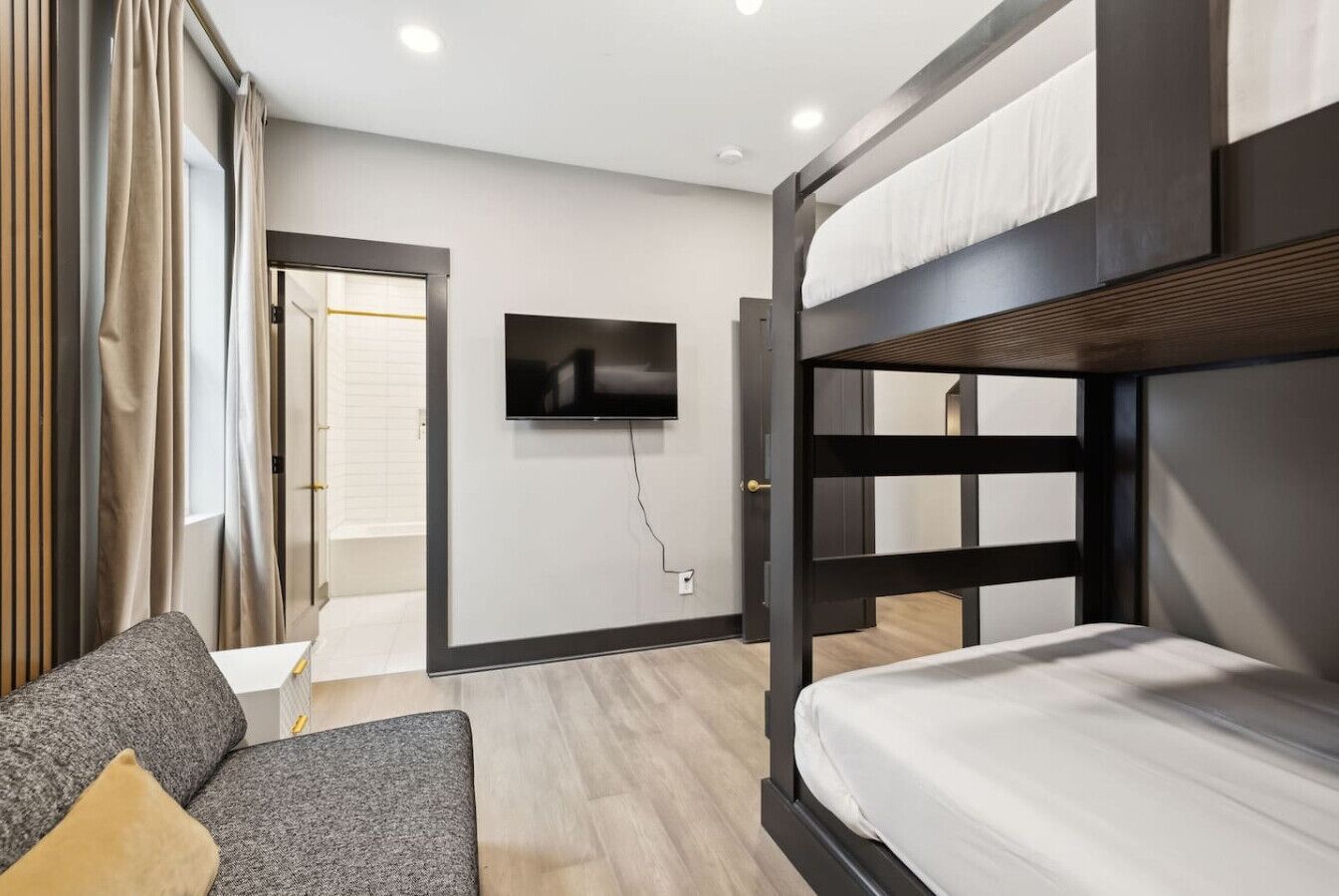 A modern hotel room with a bunk bed, a flat-screen TV mounted on the wall, a sofa with cushions, and a wooden floor.