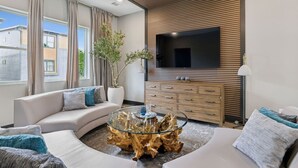 Una sala de estar moderna con un televisor de pantalla plana grande montado en una pared de paneles de madera, una mesa de centro de vidrio con base de madera natural y un sofá color beige con cojines decorativos.