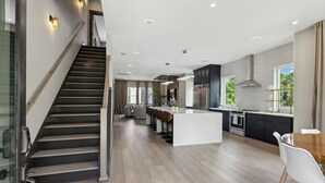 Una cocina moderna con una isla central, una escalera oscura y un comedor con sillas blancas.