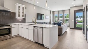 A modern kitchen with white cabinetry, a built-in oven, and a dishwasher. There is a dining area with a sofa and a TV mounted on the wall.