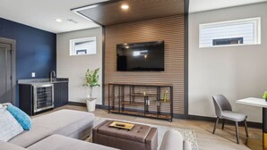 A modern living room with a sofa, a coffee table, a TV mounted on a wooden wall, a small kitchen area, and a dining table with a chair.