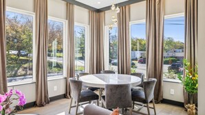A modern dining area with a round table and six chairs, large windows with curtains, and a view of the street outside.