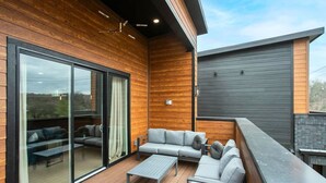 A modern outdoor living area with a wooden deck, a grey sofa set, and a glass sliding door leading to a patio. - East Nash Luxury Escape w/Rooftop Deck-Sleeps 12! (Nashville)