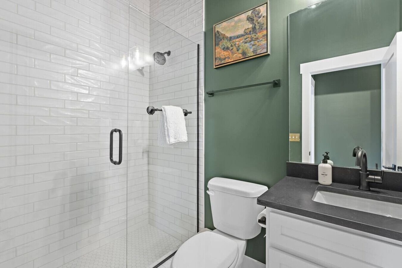 Une salle de bain moderne avec des carreaux de métro blancs, une douche en verre, des toilettes blanches, un porte-serviettes noir, un mur d’accent vert et un grand miroir avec un robinet noir.