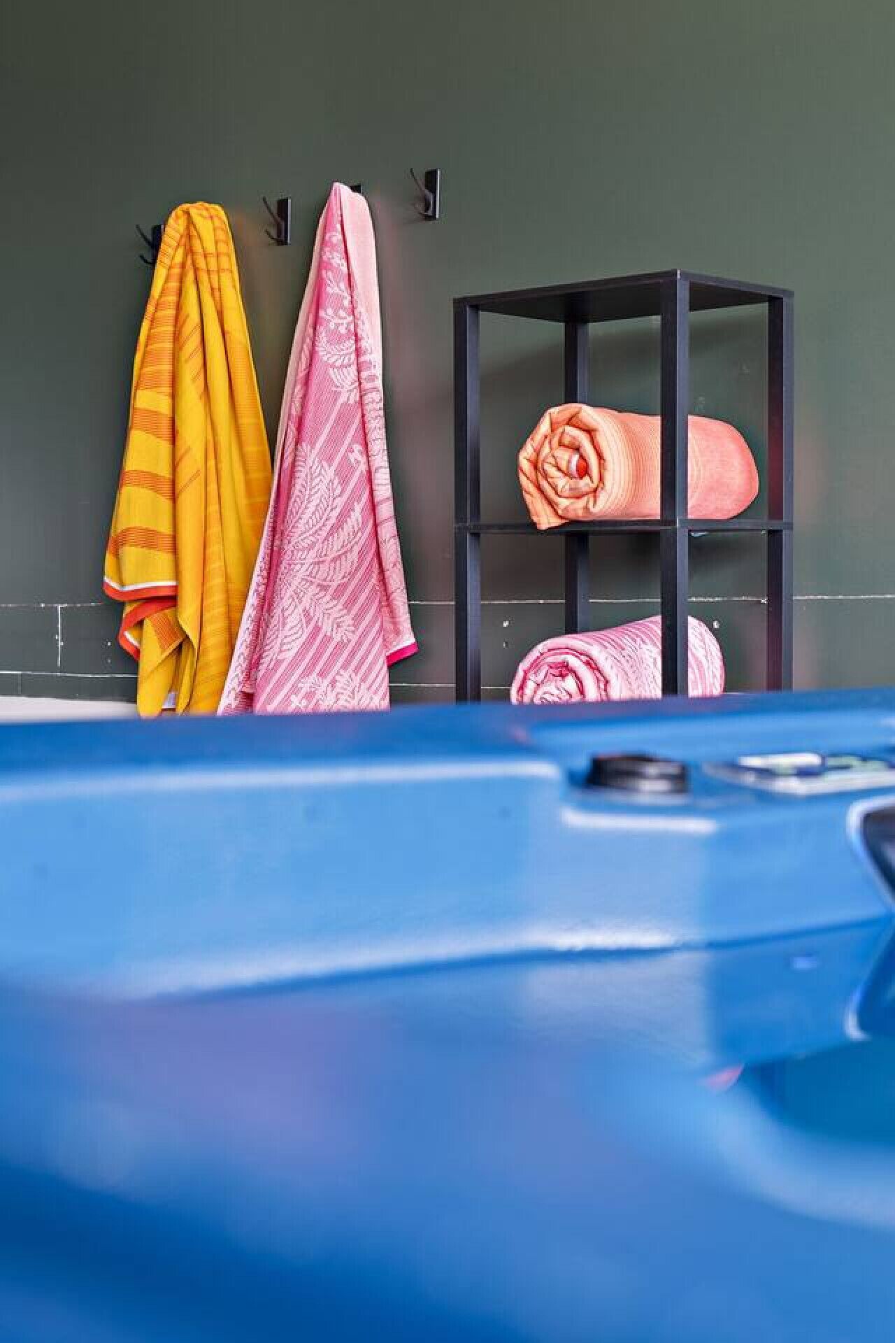 A bathroom with towels hanging on hooks and a towel rack, a blue bathtub, and a black shelf with rolled towels.