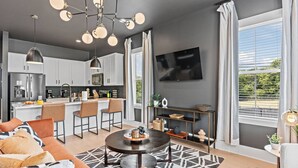 Uma sala de estar moderna com sofá, mesa de centro e área de jantar com cadeiras. Cozinha visível ao fundo, com uma TV fixada na parede.