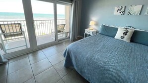 Un dormitorio con una cama grande, una mesita de noche y vista a la playa a través de una puerta corrediza de vidrio.