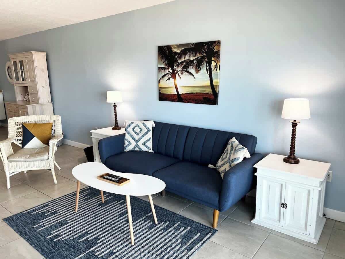 A living room with a blue sofa, white side tables, a white cabinet, and a painting of palm trees.
