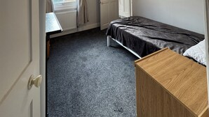 Una habitación pequeña y poco amueblada con una cama individual, una mesita de noche de madera, un radiador y una ventana con cortinas.
