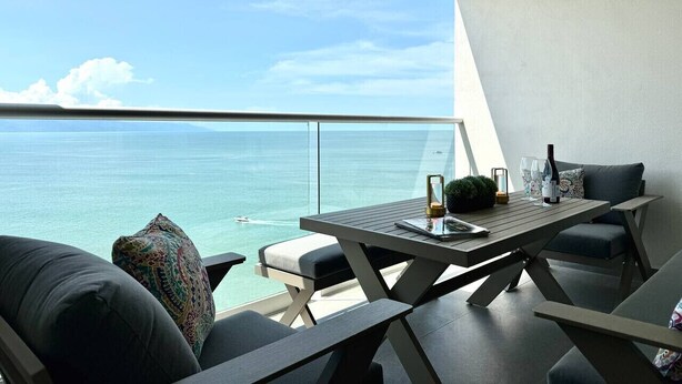 A balcony with a view of the sea, a table with chairs, and a small plant. - Moikkarentals | Beachfront 1BR Harbor171-N1702 PV (Puerto Vallarta)