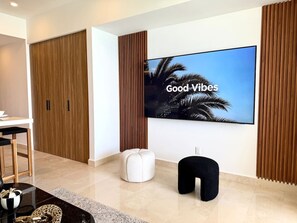 A modern living room with a large flat-screen TV, a white ottoman, a black pouf, and a wooden wardrobe. - Beachfront Harbor171 Condo | Ocean-View Balcony & Rooftop Pool (Puerto Vallarta)