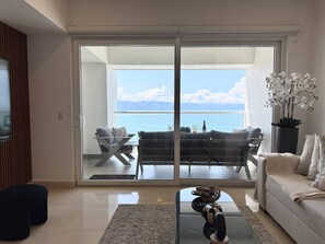 A modern living room with a glass door leading to a balcony overlooking a body of water. - Beachfront Harbor171 Condo | Ocean-View Balcony & Rooftop Pool (Puerto Vallarta)