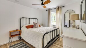 A bedroom with a bed, a bedside table, a nightstand, a ceiling fan, a window with curtains, a mirror, and a lamp. - Direct Lakefront Retreat | Indian Ranch | Casinos (Thompson)