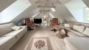 Een ruime zolderkamer met hoge plafonds, houten balken en dakramen. Er staat een grote bank, twee fauteuils, een klein rond tafeltje en een televisie.