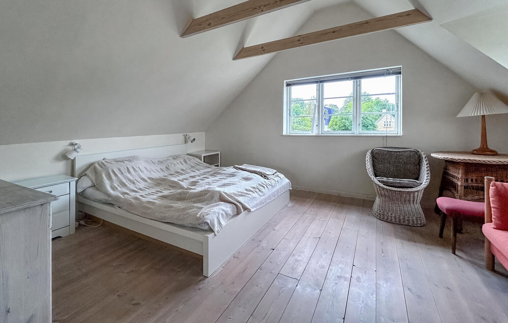 A bedroom with a single bed, a chair, a small table, and a window with a view of greenery.