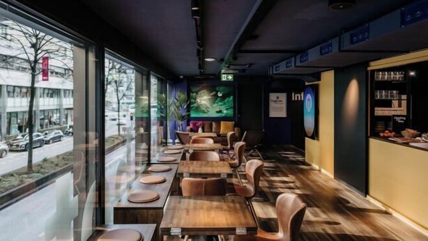 Ein modernes Restaurant mit Holztischen und -stühlen, großen Fenstern mit Blick auf die Straße und einem Neonzeichen an der Wand.