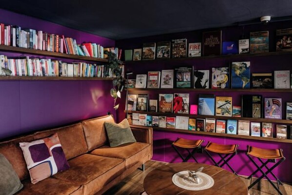 A room with a purple wall, a brown sofa, a coffee table, and a bookshelf filled with books and framed pictures. - Capsule Hotel - Lucerne Old Town (Lucerne)
