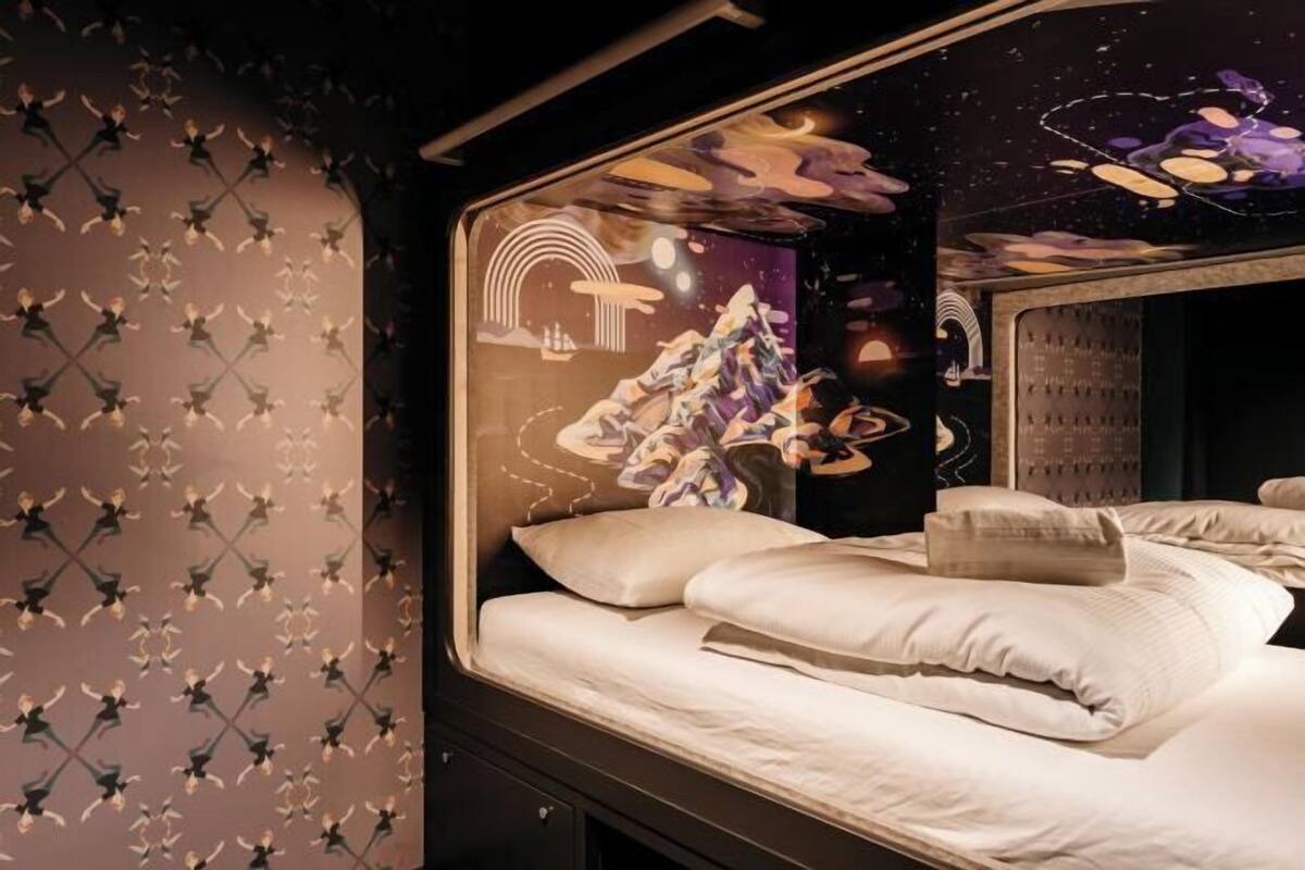 Een bed met witte lakens, een donkere muur met een geometrisch patroon en een groot raam met een kleurrijke abstracte muurschildering.
