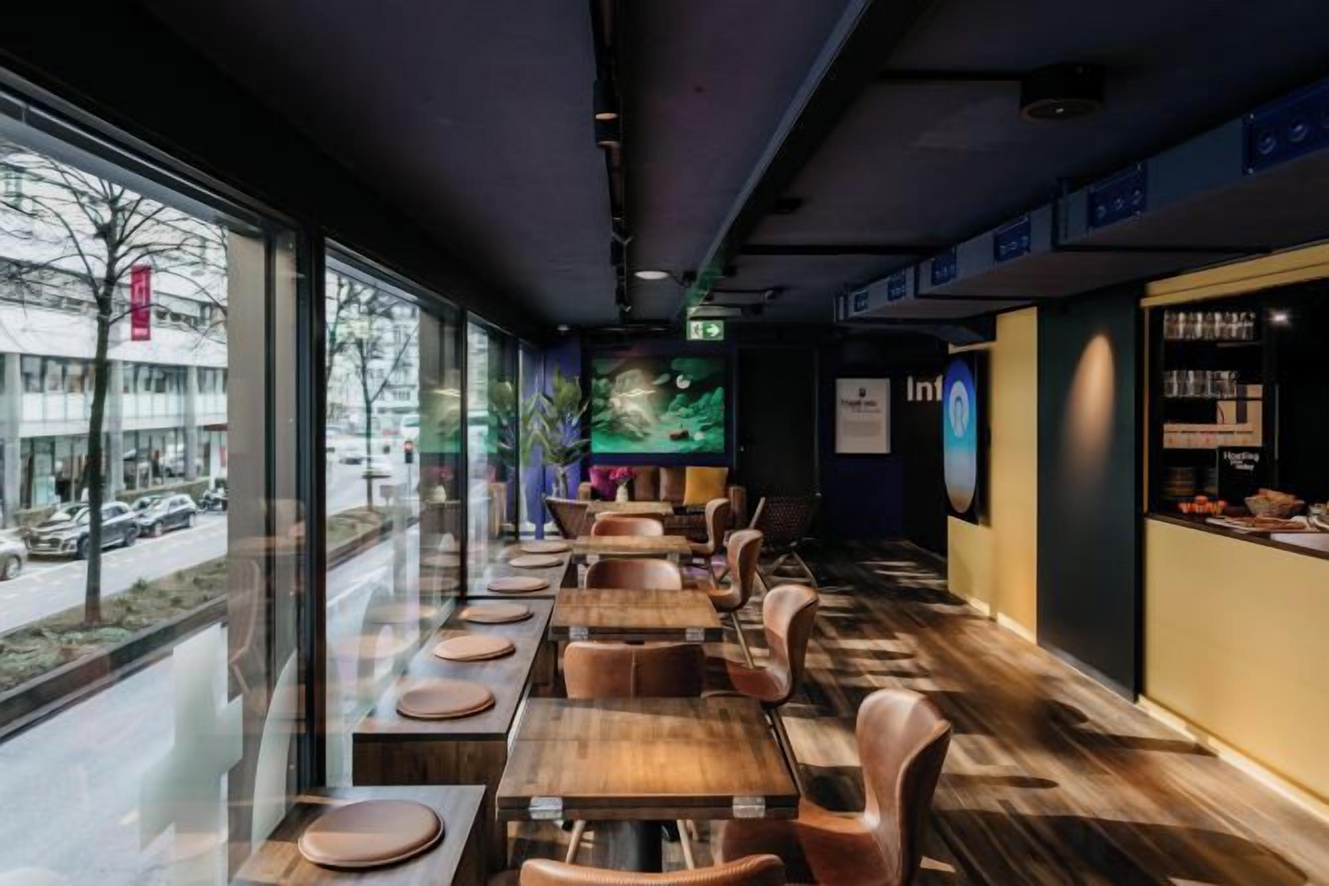 A restaurant with long tables and chairs, a view of the street outside, and a menu display on the right.