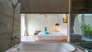 Un bagno moderno con una vasca da bagno ampia, una doccia e vista su una camera da letto con un letto e un comodino.