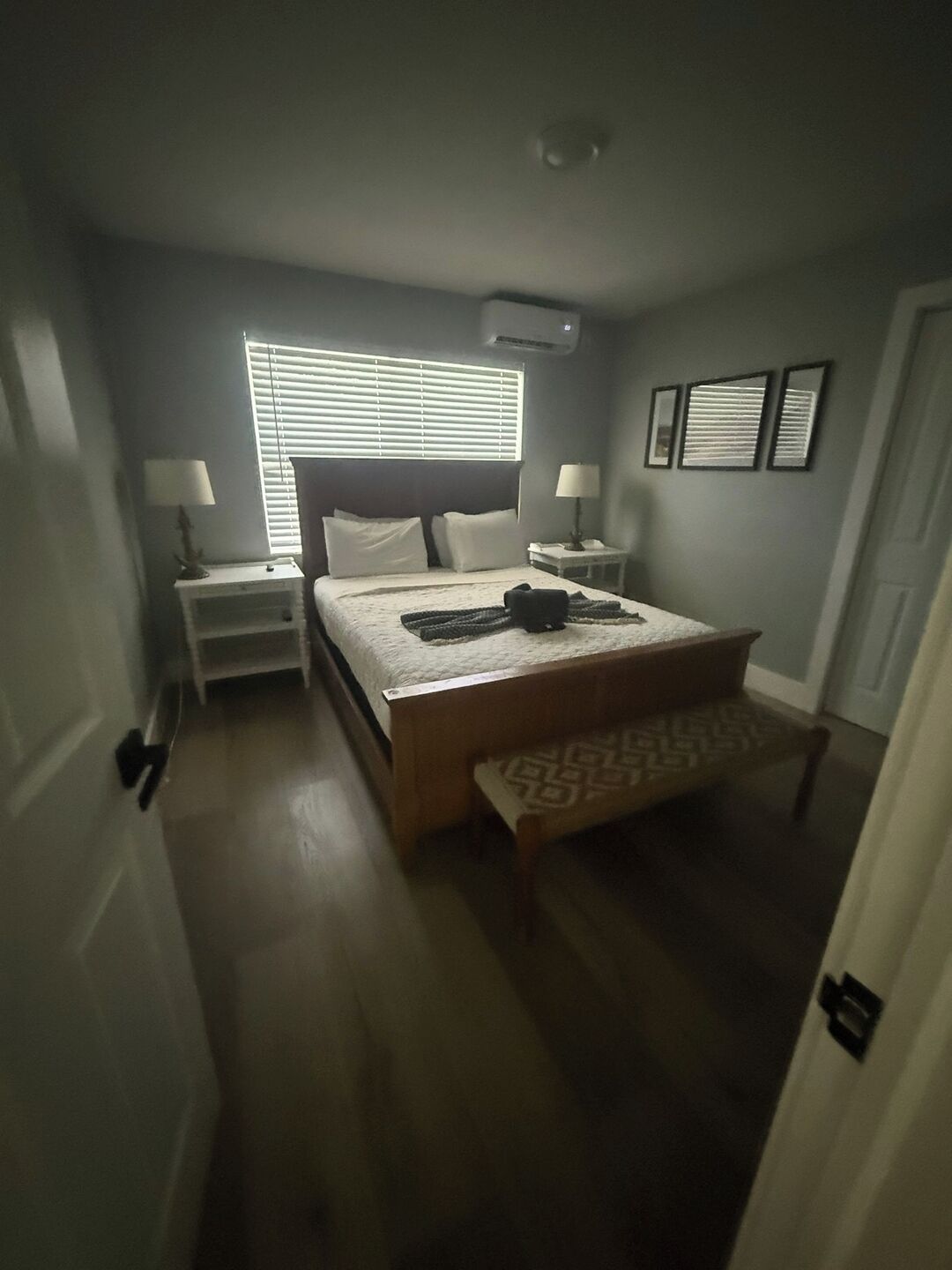 A bedroom with a bed, bedside tables, a bench, a window with blinds, and wall-mounted pictures.