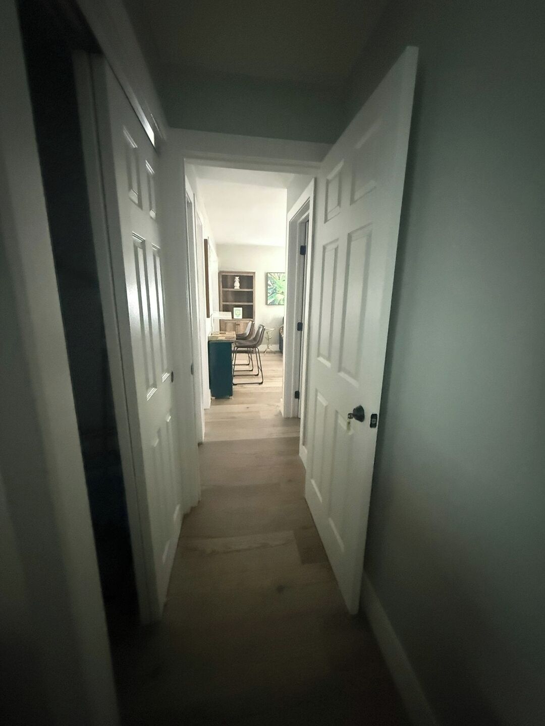 A narrow hallway with white doors leading to a room with a wooden floor and a dining area in the background.