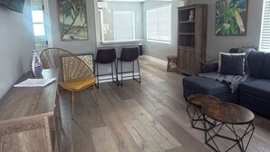 Amplia sala de estar con suelo de madera, un sofá, una mesa de centro y dos sillones. Hay dos ventanas con persianas, una estantería y un televisor de pantalla plana montado en la pared.