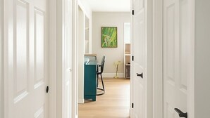 Un pasillo con puertas blancas que dan a una habitación con un escritorio, una silla, y un cuadro en la pared.
