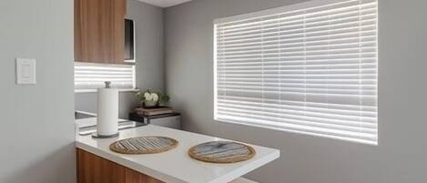 A modern kitchen with a white countertop, two red stools, a wooden cabinet, and a window with blinds.