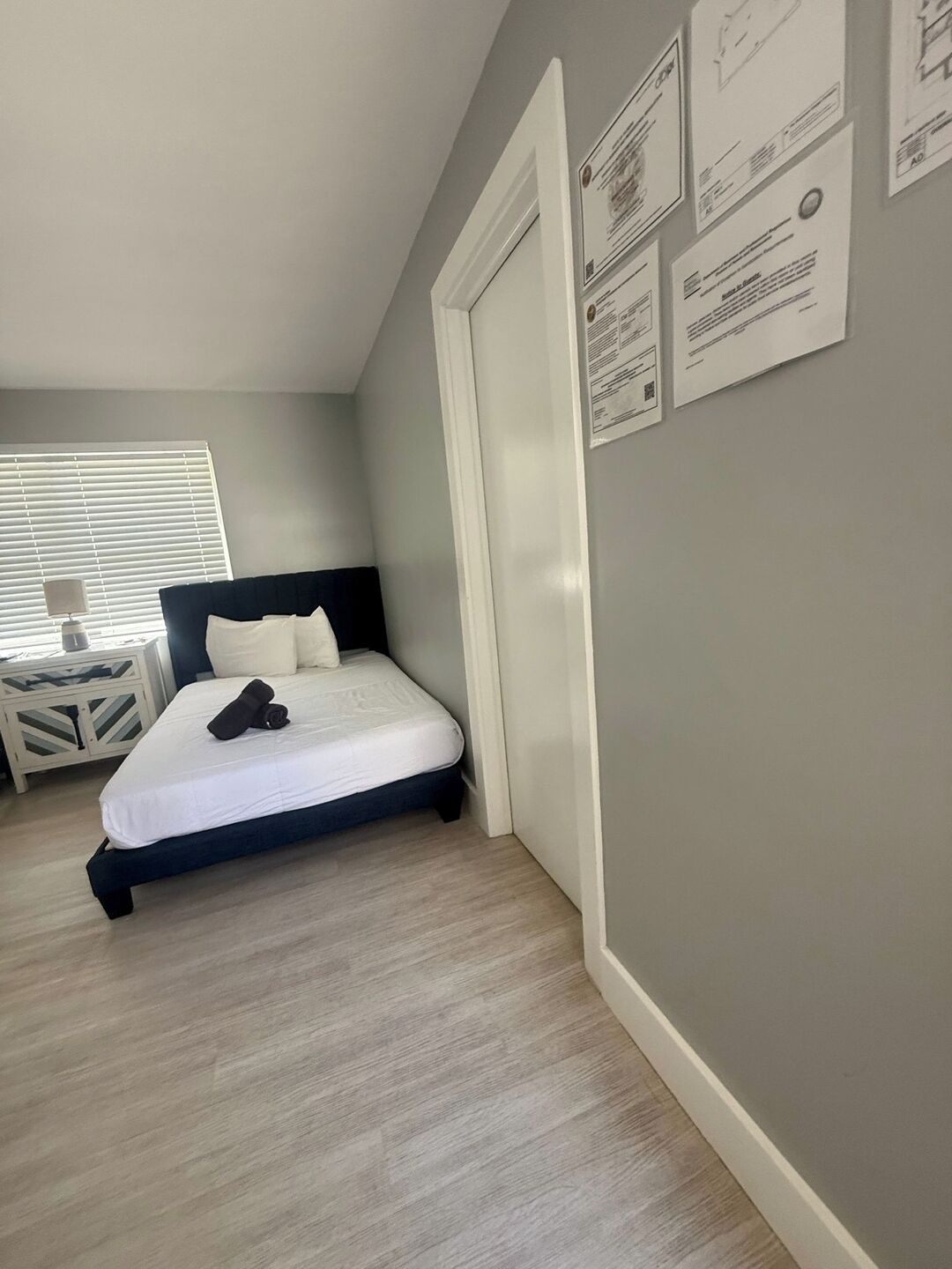 A bedroom with a bed, a nightstand, a window with blinds, and a door.