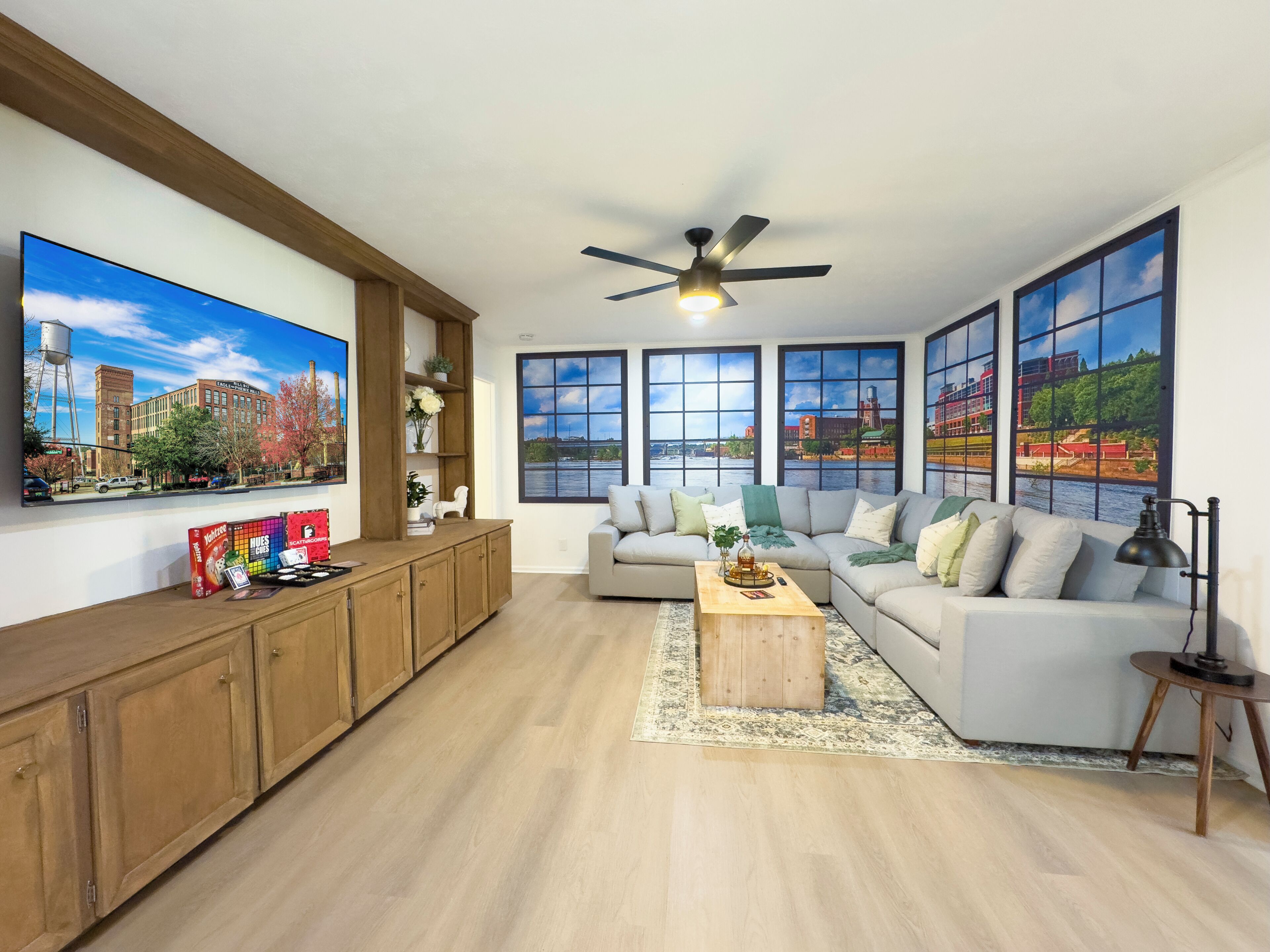 A spacious living room with a large flat-screen TV, a ceiling fan, and large windows with city views.